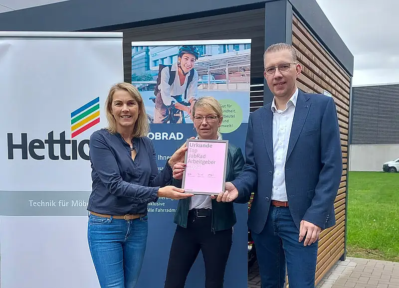 Monika Titze, von der Hettich Gruppe, steht zusammen mit einer Frau und einem Mann vor einem Poster mit dem Schriftzug Hettich und dem Hettich Logo. Sie alle tragen blaue Geschäftskleidung Monika Titze, von der Hettich Gruppe, steht zusammen mit einer Frau und einem Mann vor einem Poster mit dem Schriftzug Hettich und dem Hettich Logo. Sie alle tragen blaue Geschäftskleidung